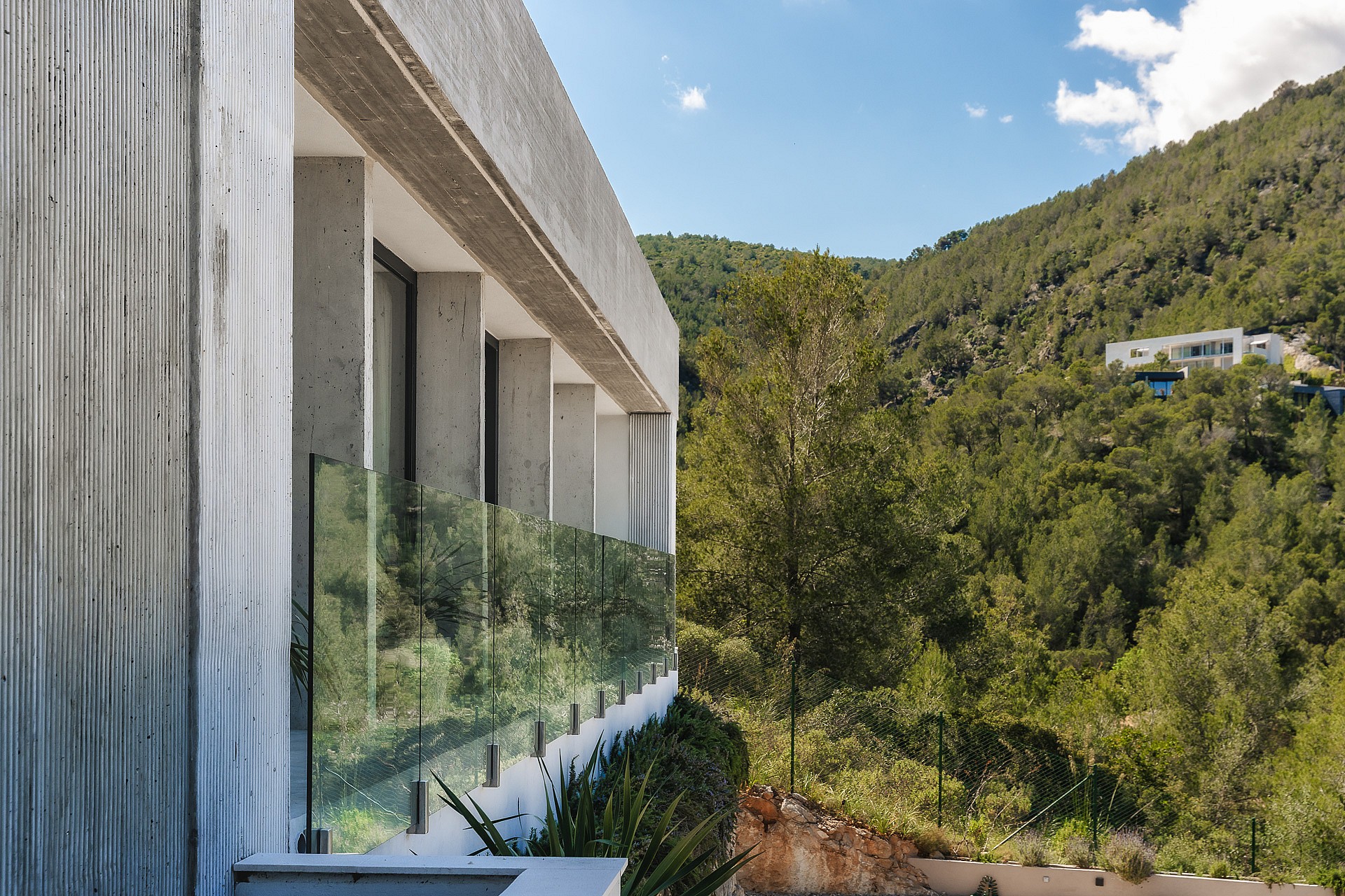 Side profile of a textured concrete villa exterior with glass railings overlooking green hills under a blue sky.