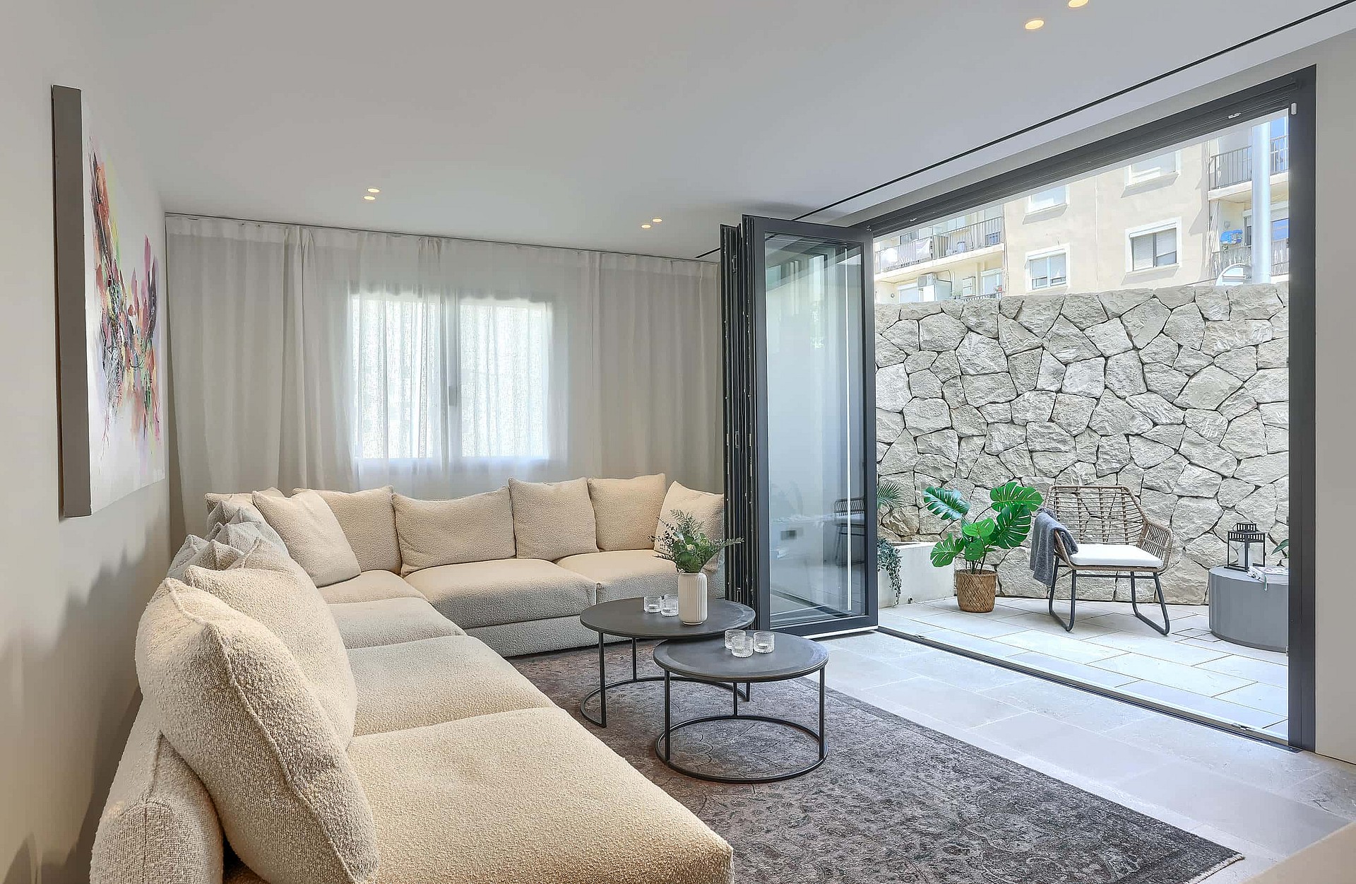 A contemporary living room with a large cream sectional sofa, dark round coffee tables, and floor-to-ceiling glass doors opening to a stone-walled courtyard.