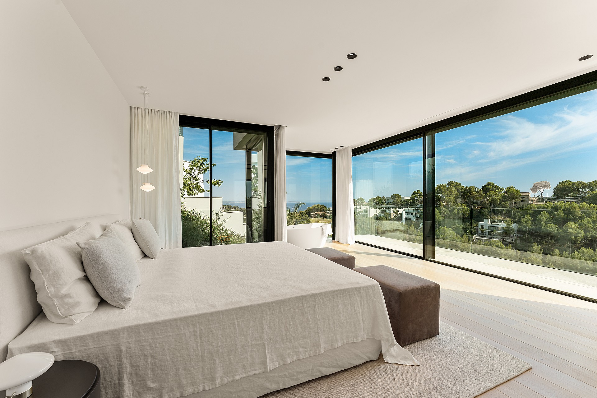 A modern master bedroom with floor-to-ceiling glass walls overlooking a wooded landscape and a distant horizon line.