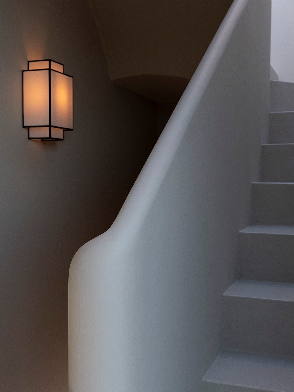 A curved plaster staircase with a modern black lantern wall sconce emitting a warm glow in a minimalist hallway.