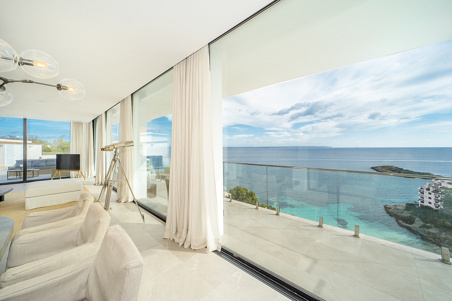 Modern living room with floor-to-ceiling sliding glass doors opening to a terrace, light beige furniture, and sea views.