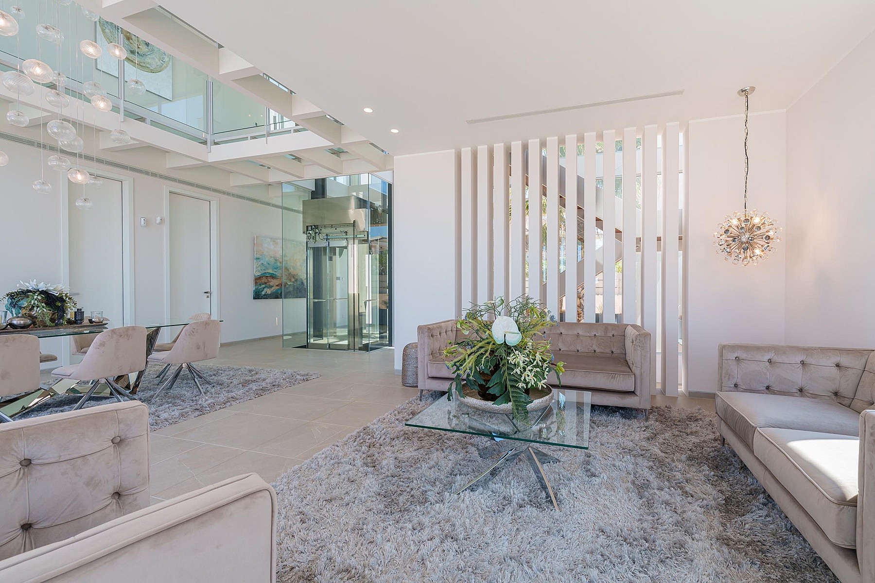 Contemporary living room featuring a central glass elevator, light beige sofas, a textured area rug, and vertical slat partition.