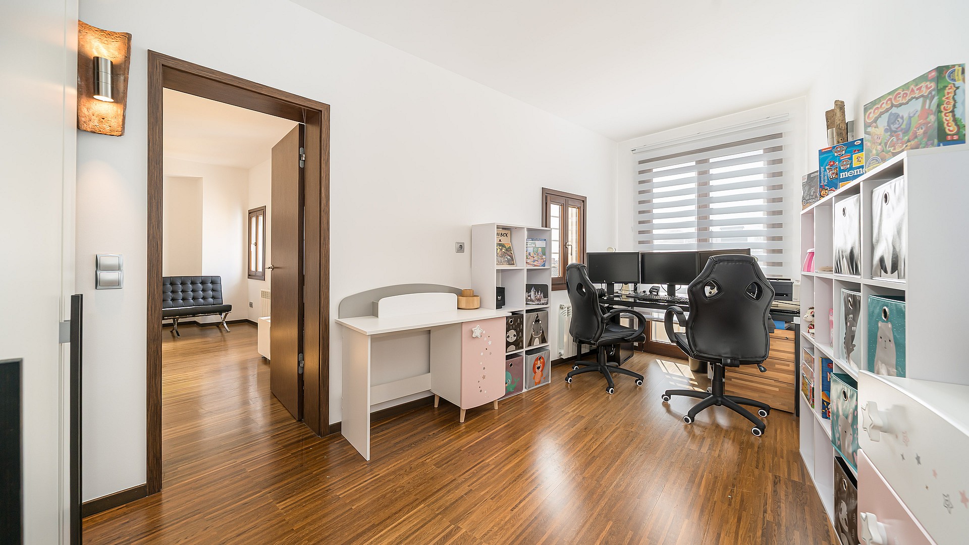 A bright room with dark wood floors, a desk with multiple monitors, and white storage shelving units.