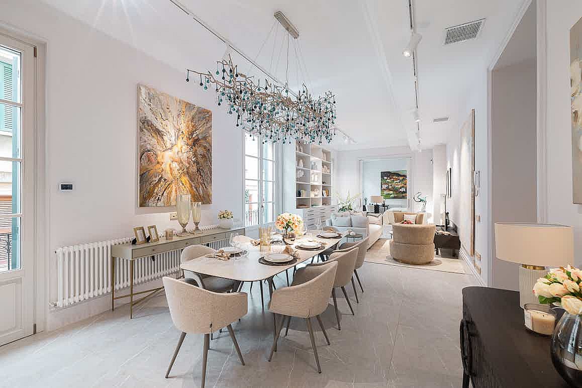 Dining area with a rectangular light-wood table, cream upholstered chairs, and a large sculptural chandelier overhead.