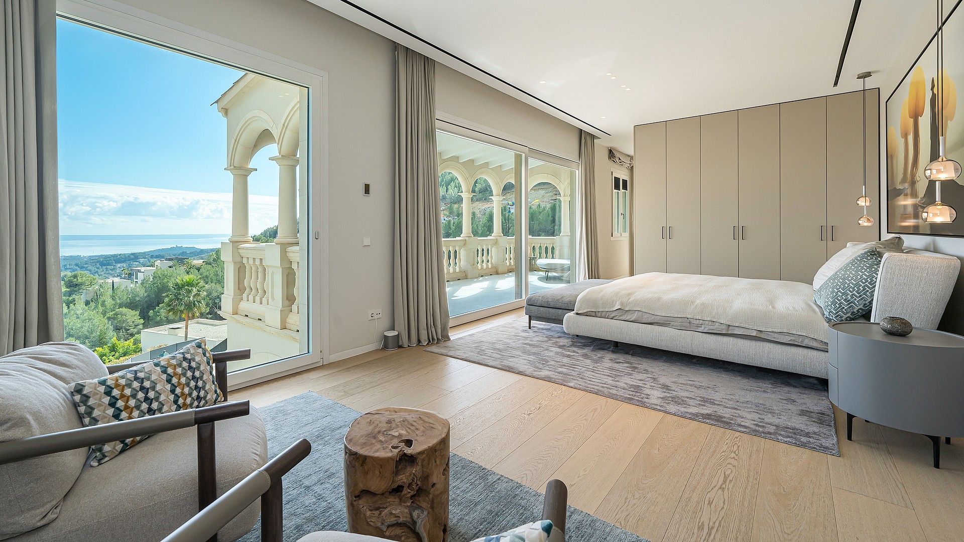 A spacious bedroom with a king-sized bed, soft grey carpet, floor-to-ceiling glass doors opening to a stone-railed balcony.