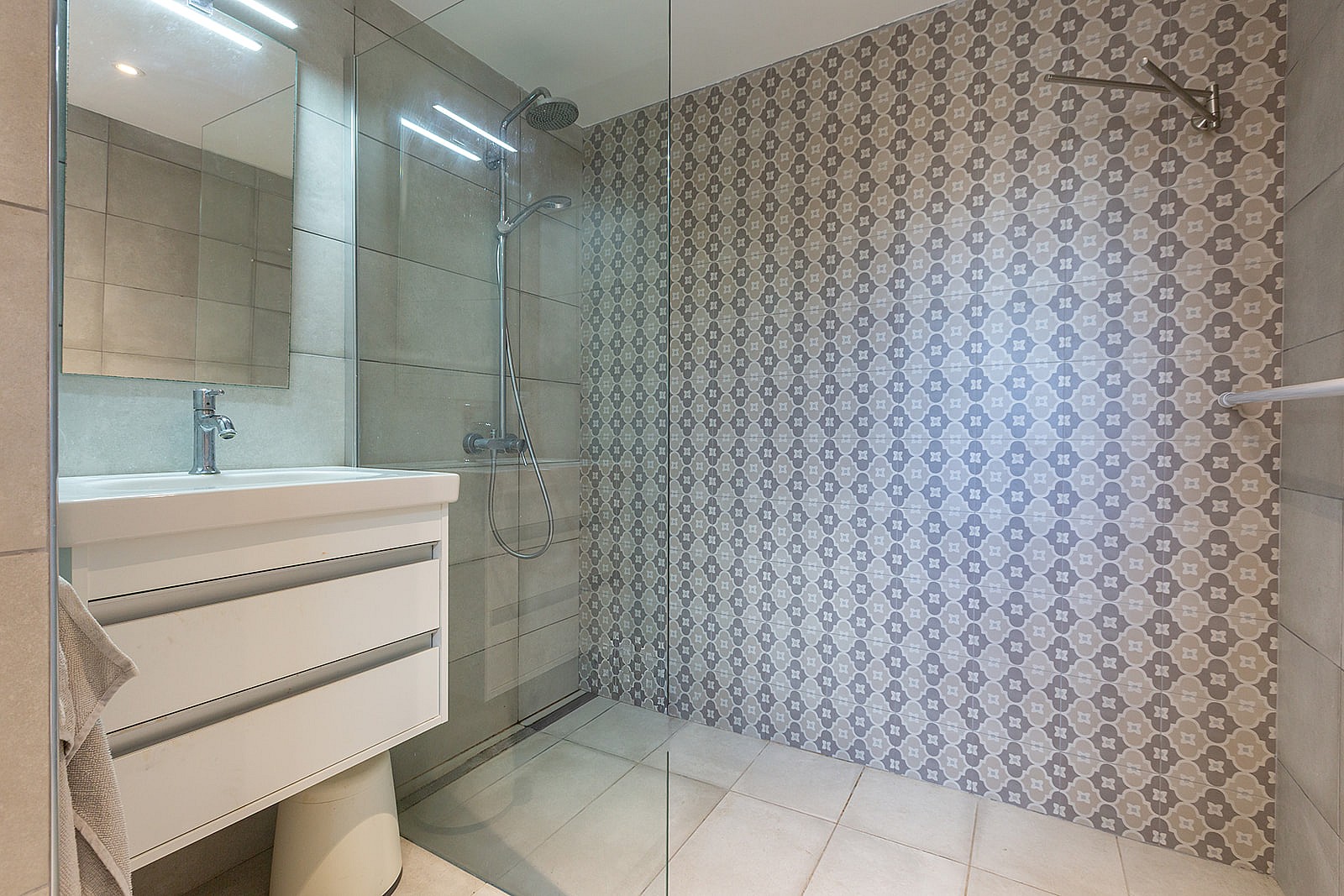 A modern bathroom featuring a white vanity, a large glass-enclosed shower with decorative geometric tilework, and neutral stone floors.