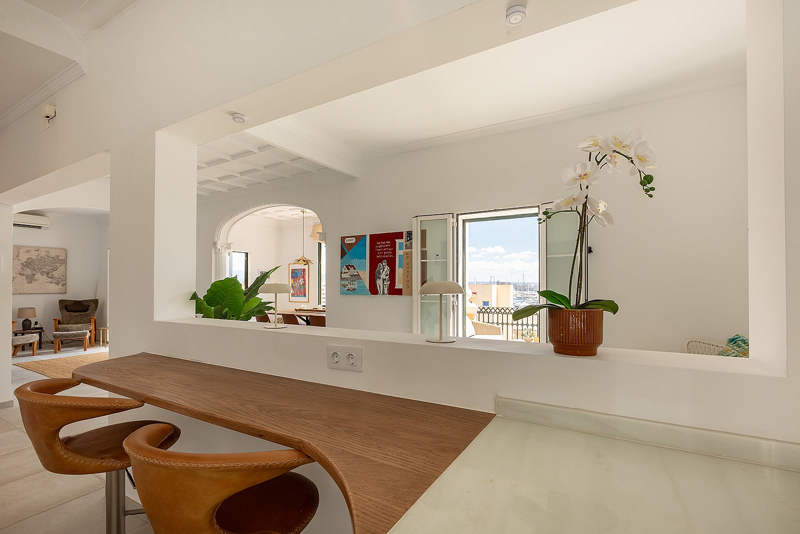 A white kitchen breakfast bar with wooden countertop and brown leather stools looking through an arched opening to a room.