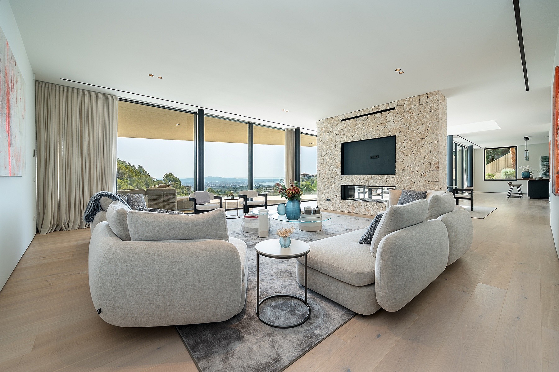 Contemporary living room with light oak floors, a textured stone fireplace wall, and floor-to-ceiling windows facing the sea.