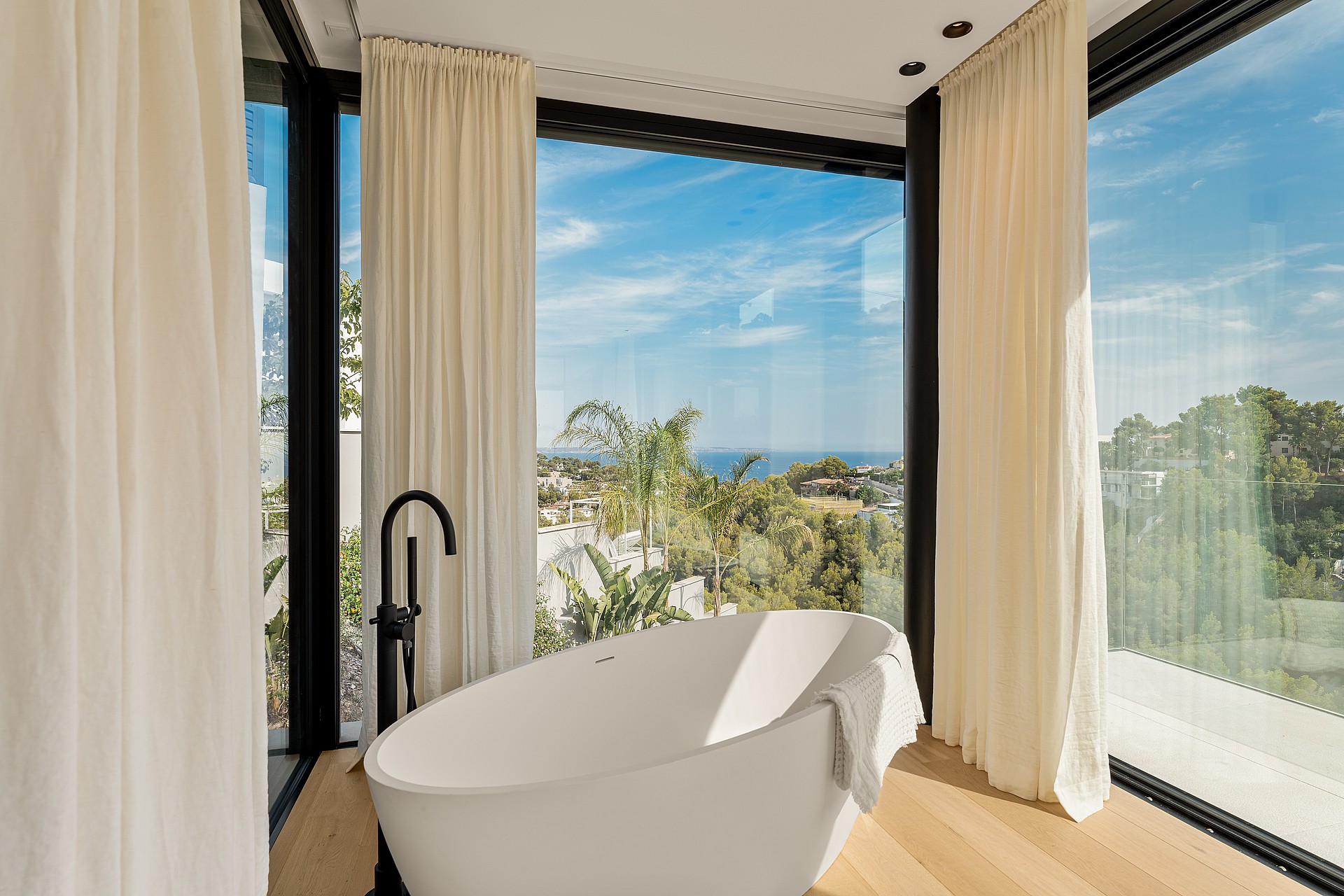 Freestanding white bathtub in a bathroom with floor-to-ceiling windows, cream curtains, and light wood flooring.