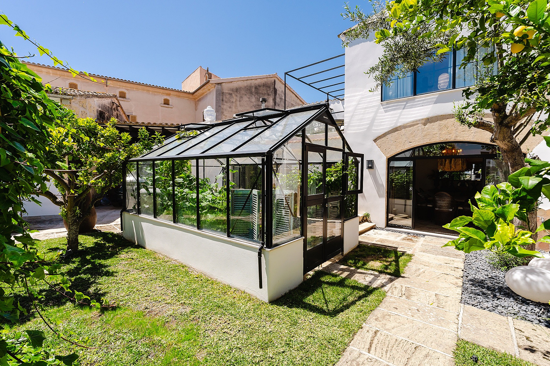 A glass and black metal greenhouse located in a private landscaped garden with a stone path and lemon tree.