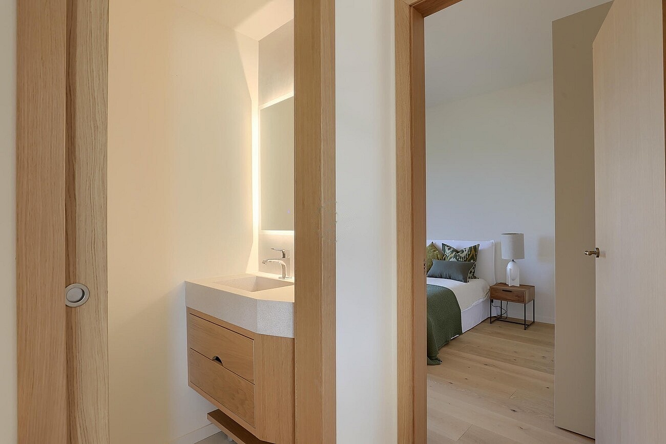 A view through a wooden door frame into a bedroom with a view of a minimalist wooden vanity unit in the foreground.