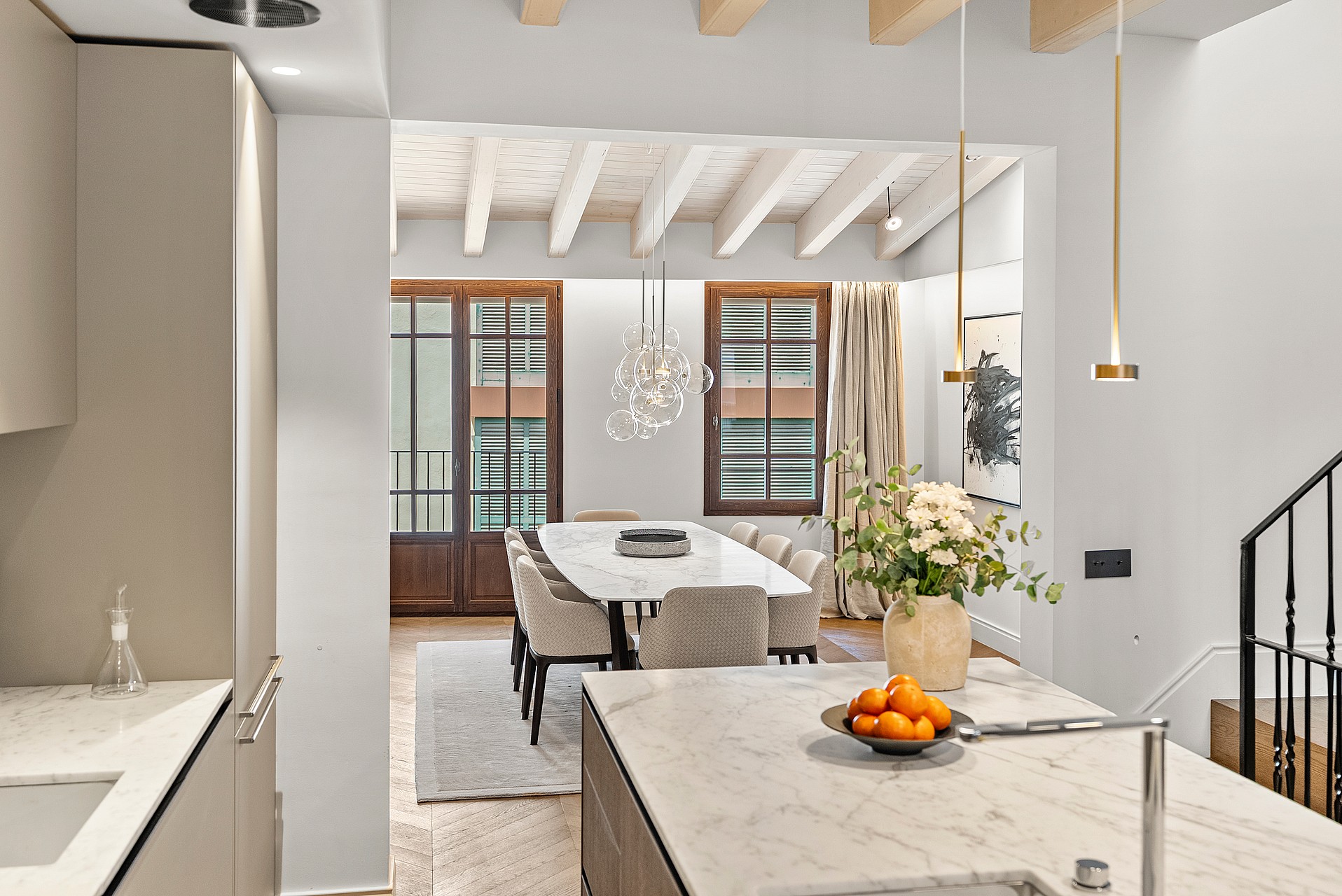 Kitchen island view to dining area featuring marble tables, exposed wood beams, grey chairs, and a wrought iron staircase.