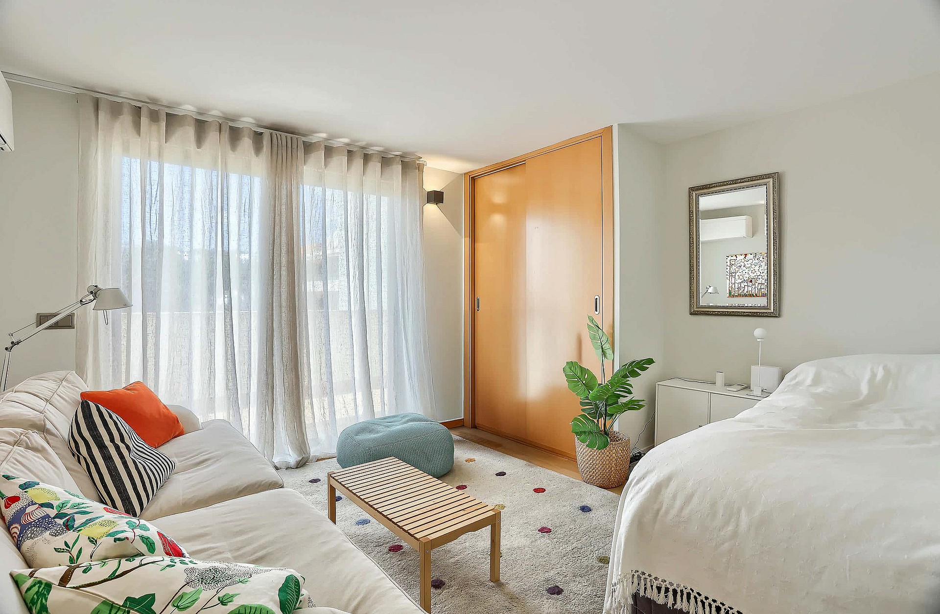 A bedroom with a double bed, large wooden floor-to-ceiling wardrobe, sheer white curtains, and a patterned area rug.