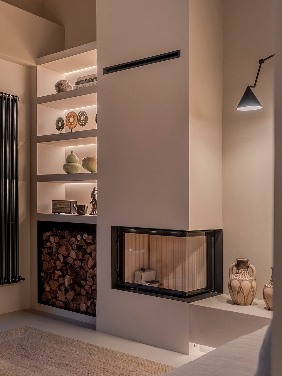 Modern glass fireplace with recessed LED shelving, stacked wood storage, and a minimalist black wall lamp.