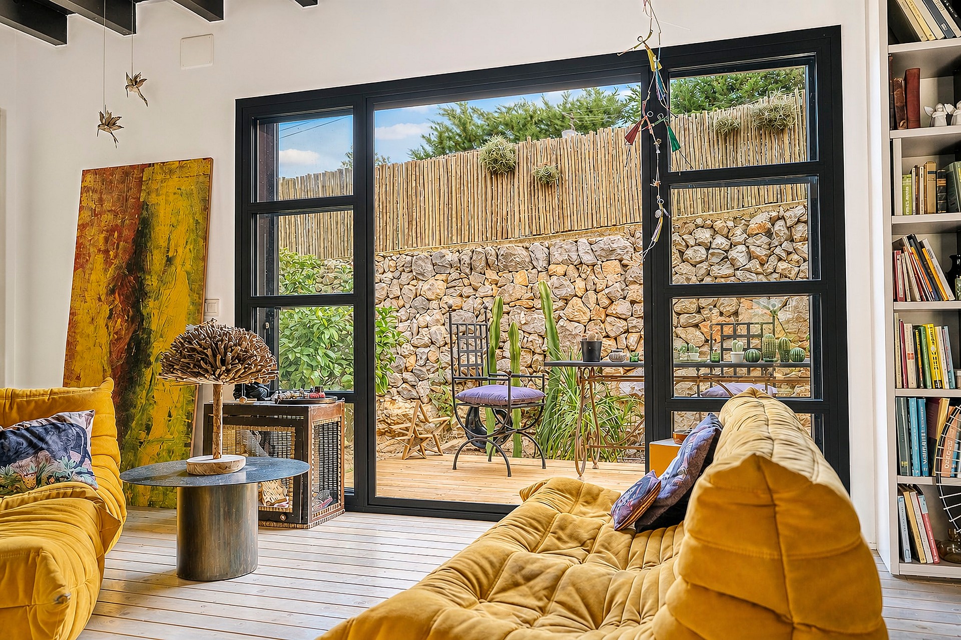 Interior living room with yellow modular sofa, glass coffee table, and large black-framed doors opening to a stone garden.