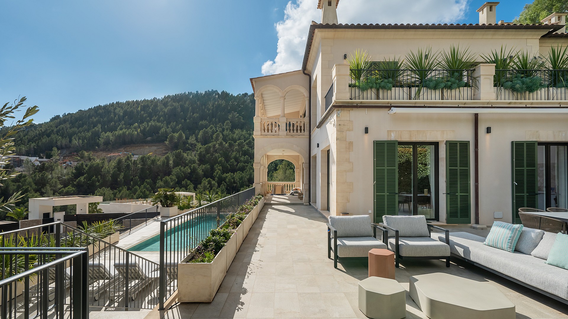 Exterior facade of a cream stone villa with a long swimming pool and shaded outdoor seating area overlooking hills.