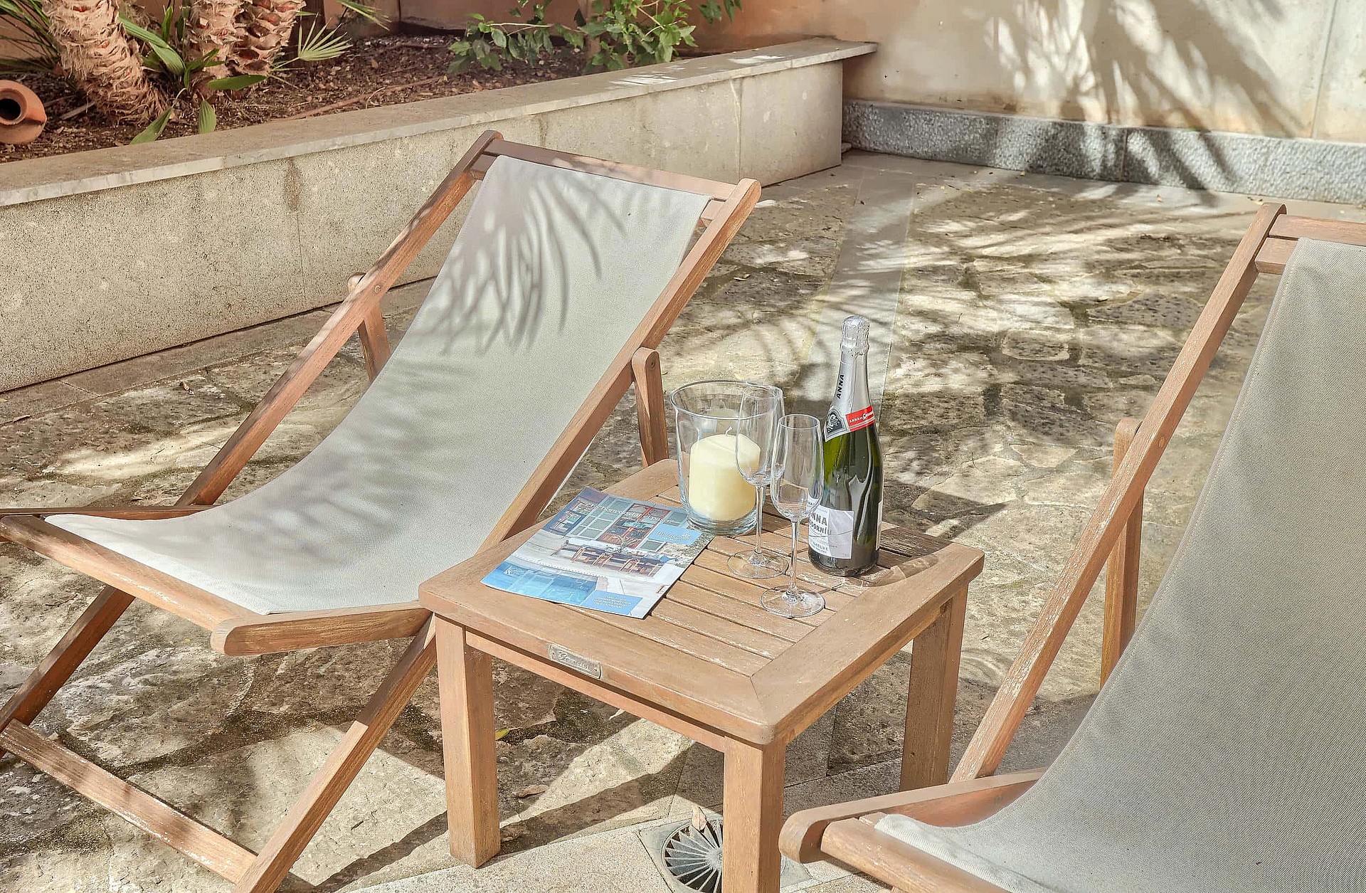 Two wooden lounge chairs with grey fabric seats arranged on a stone patio beside a small square side table with drinks.