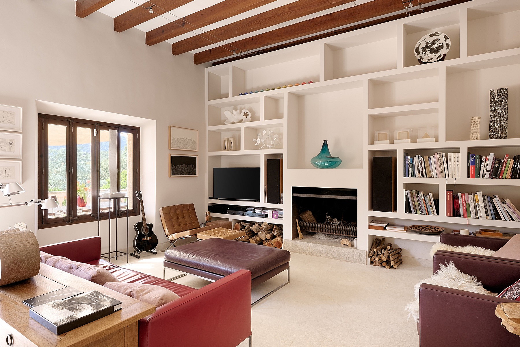 Living room with exposed wood ceiling beams, white built-in shelving unit, dark red sofa, and an open fireplace.