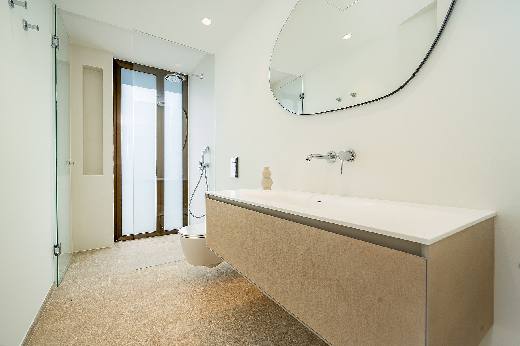 Bright bathroom with a floating beige vanity, white countertop, irregular shaped mirror, and glass-enclosed shower area.