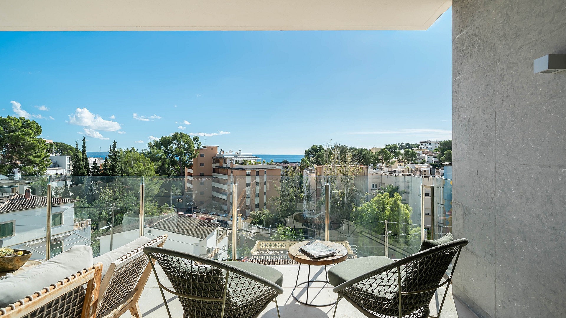 A balcony view featuring two dark wicker armchairs and a small round wooden table overlooking a coastal town and the sea.