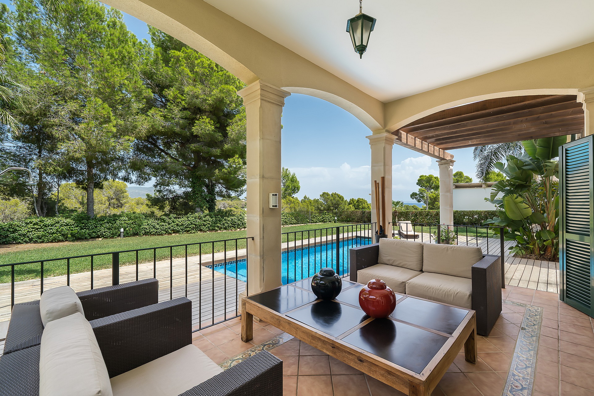 A covered terrace with comfortable outdoor seating overlooks a blue swimming pool and surrounding green gardens on a sunny day.
