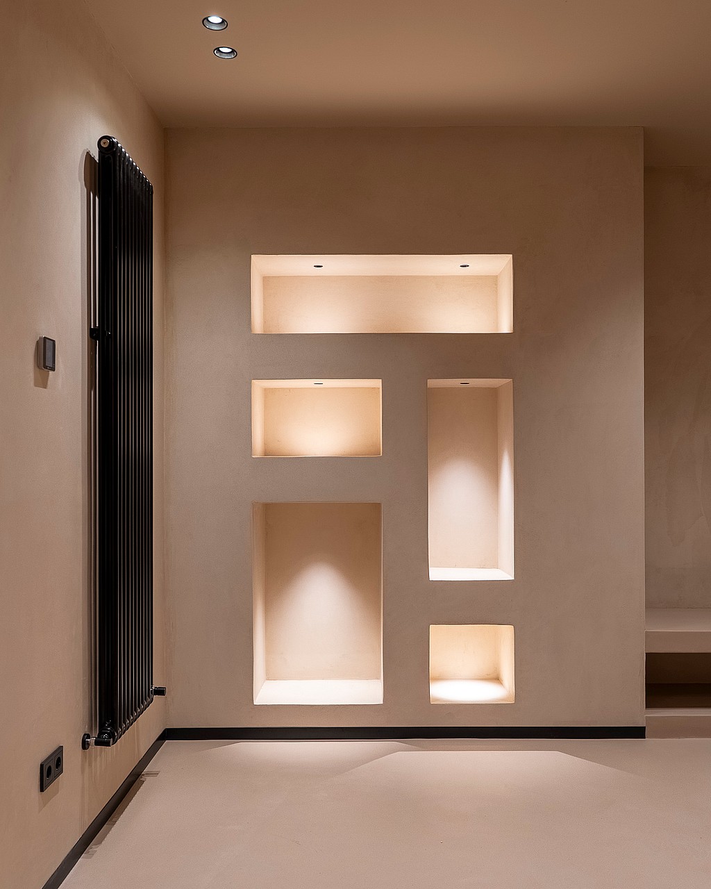 A smooth beige interior wall with four asymmetrical recessed alcoves, each illuminated by warm integrated spotlights, next to a black vertical radiator.