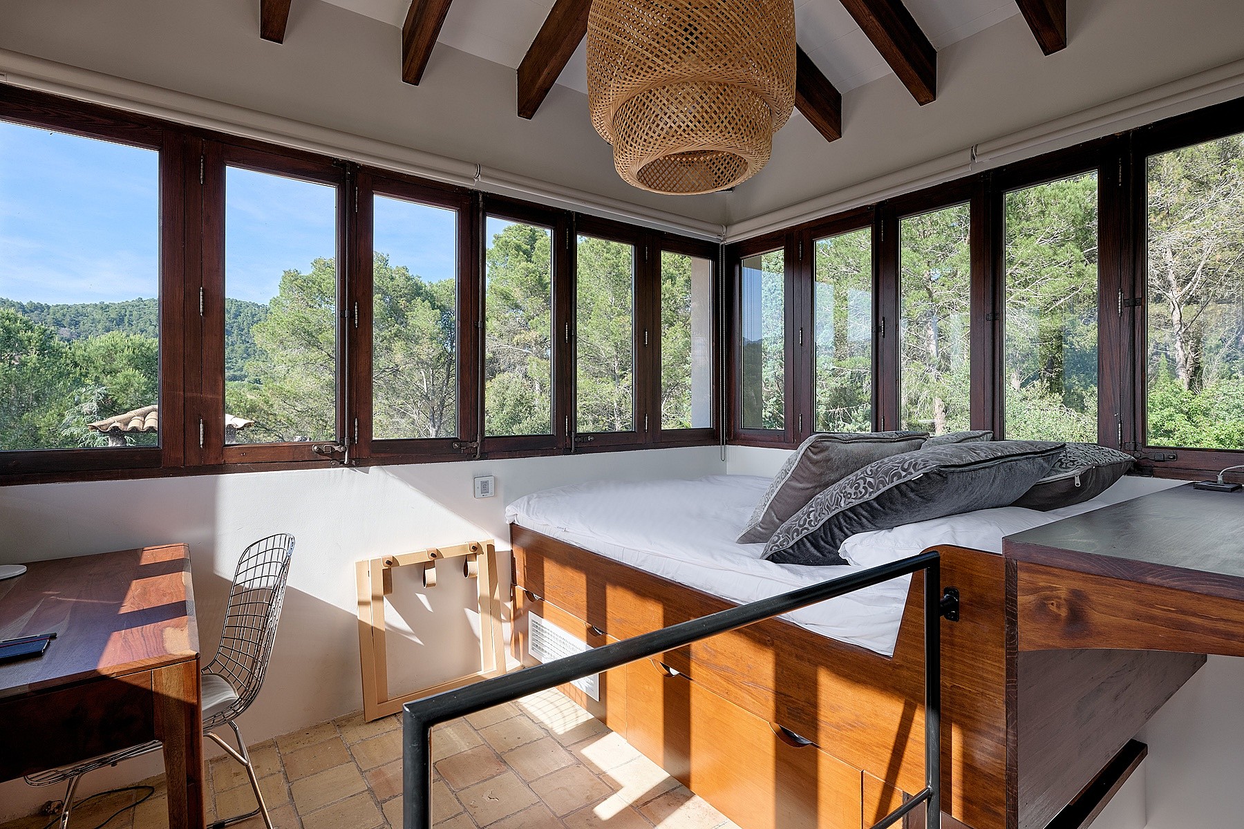 A sunlit bedroom in a finca with wooden ceiling beams, large windows overlooking forest, and a built-in wooden bed frame.