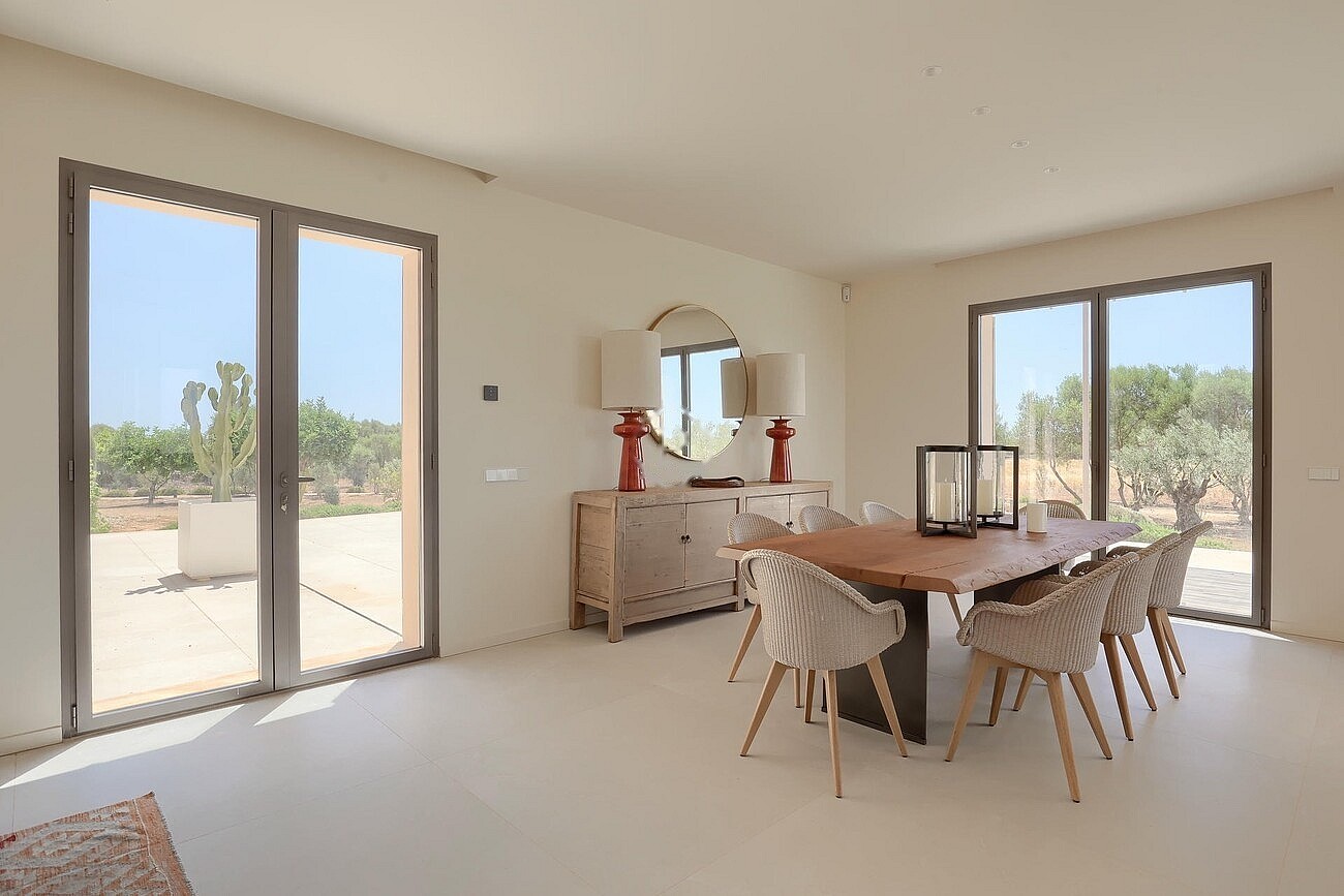 Dining room featuring a wooden table, upholstered chairs, a sideboard with lamps, and large glass doors opening to a garden.