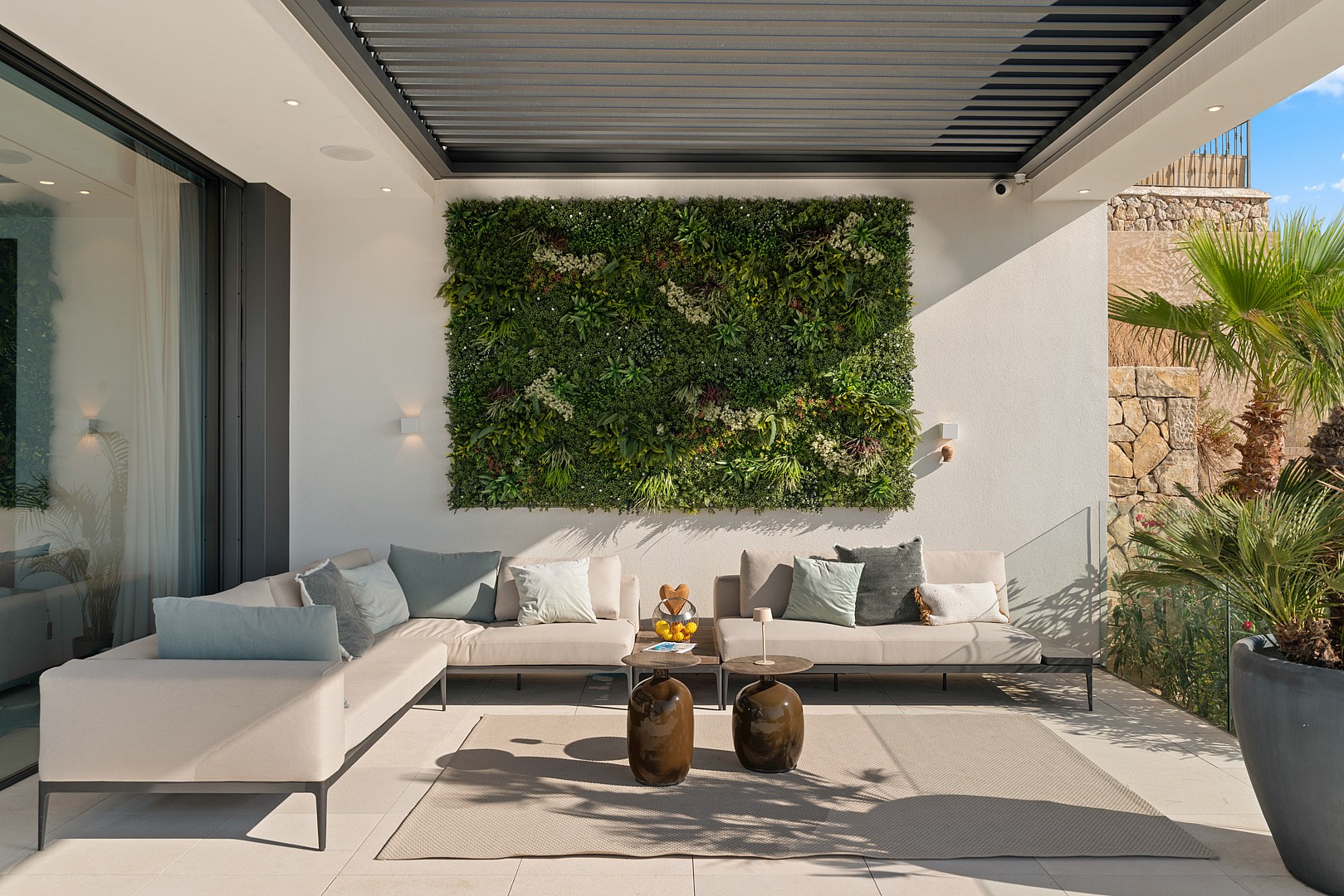 A shaded outdoor lounge area featuring a large vertical garden wall, cream-colored modular sofas, and stone side tables.