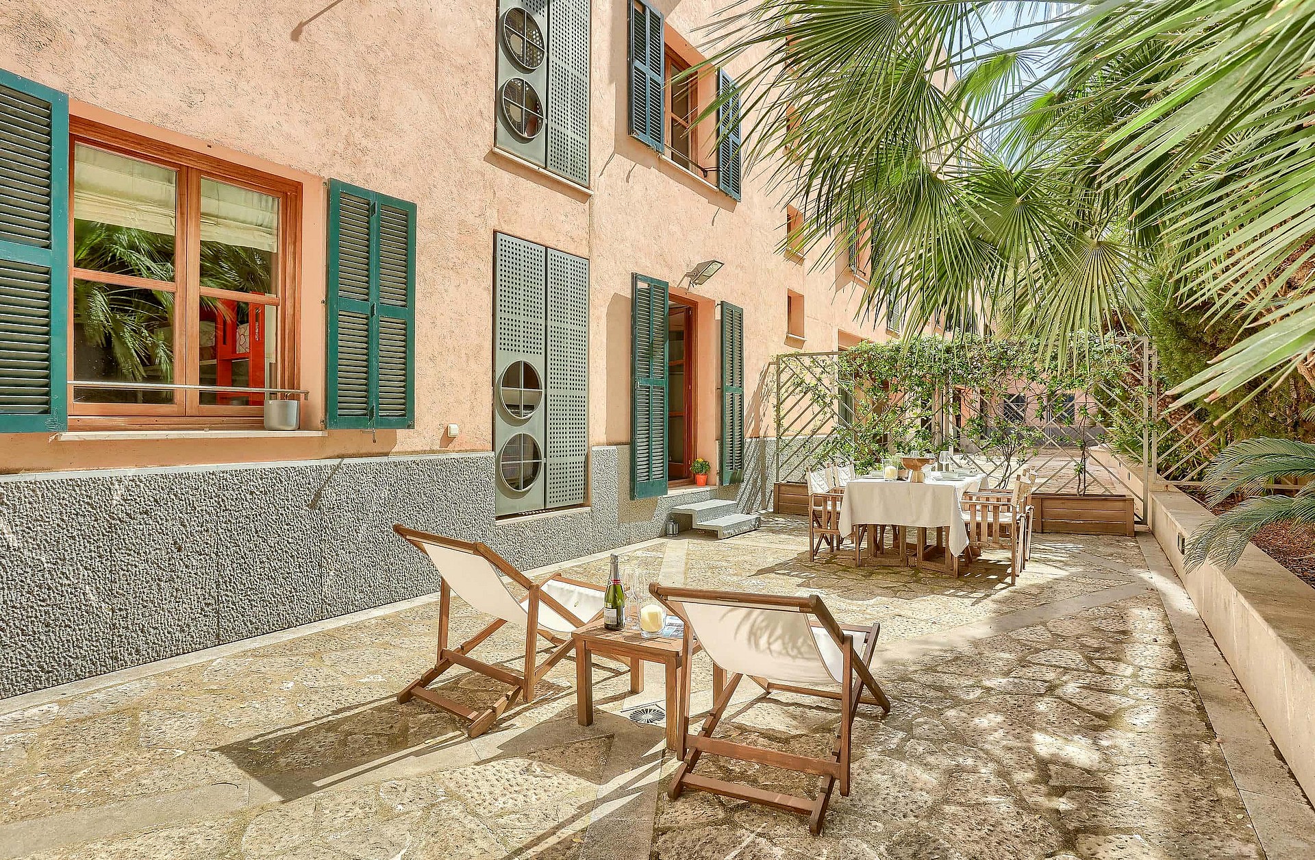 A private outdoor terrace in the Old Town featuring gravel flooring, white folding lounge chairs, a dining set, and pink stucco walls with green shutters.
