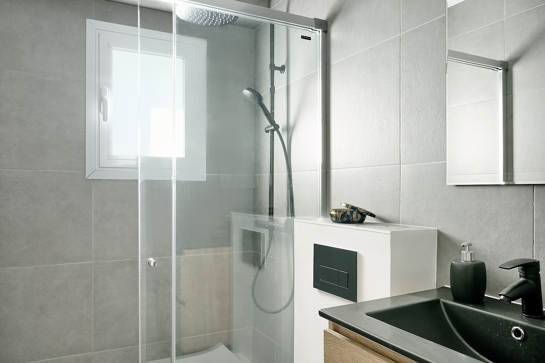 A bathroom with grey tiled walls, a glass-enclosed shower with a rainfall head, and a vanity with a black sink.