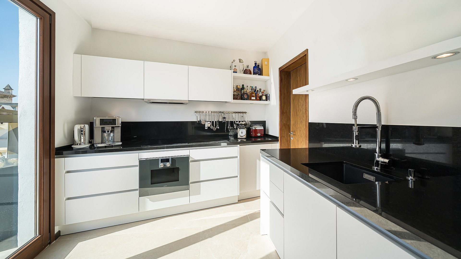 Bright modern kitchen with white handleless cabinets, black granite worktops, and stainless steel Gaggenau appliances.