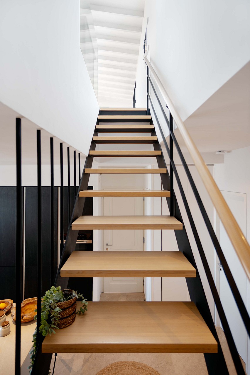 A modern, open-tread staircase featuring light oak steps and a sleek black metal railing, ascending toward a bright landing.
