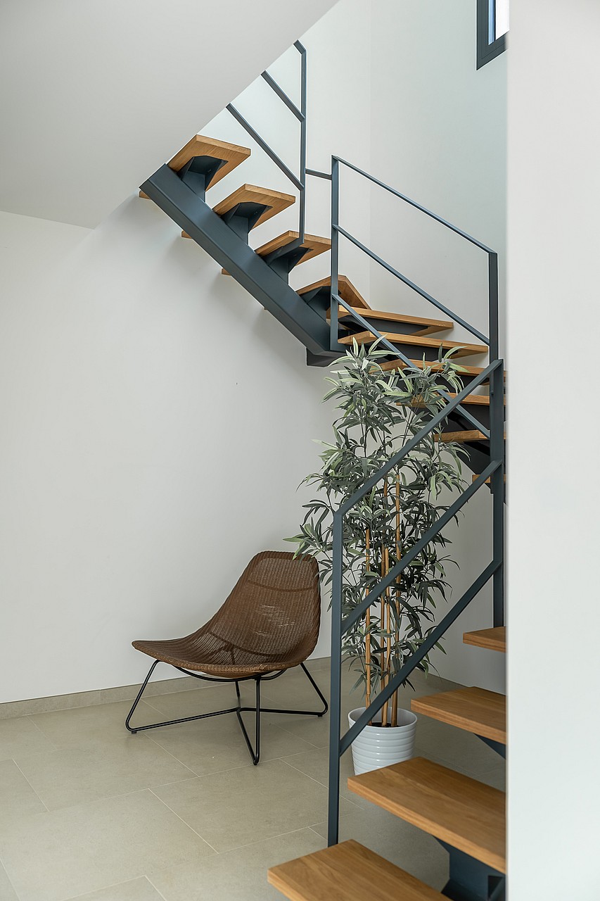A modern staircase with dark metal frame, oak steps, and a white interior, featuring a woven accent chair underneath.