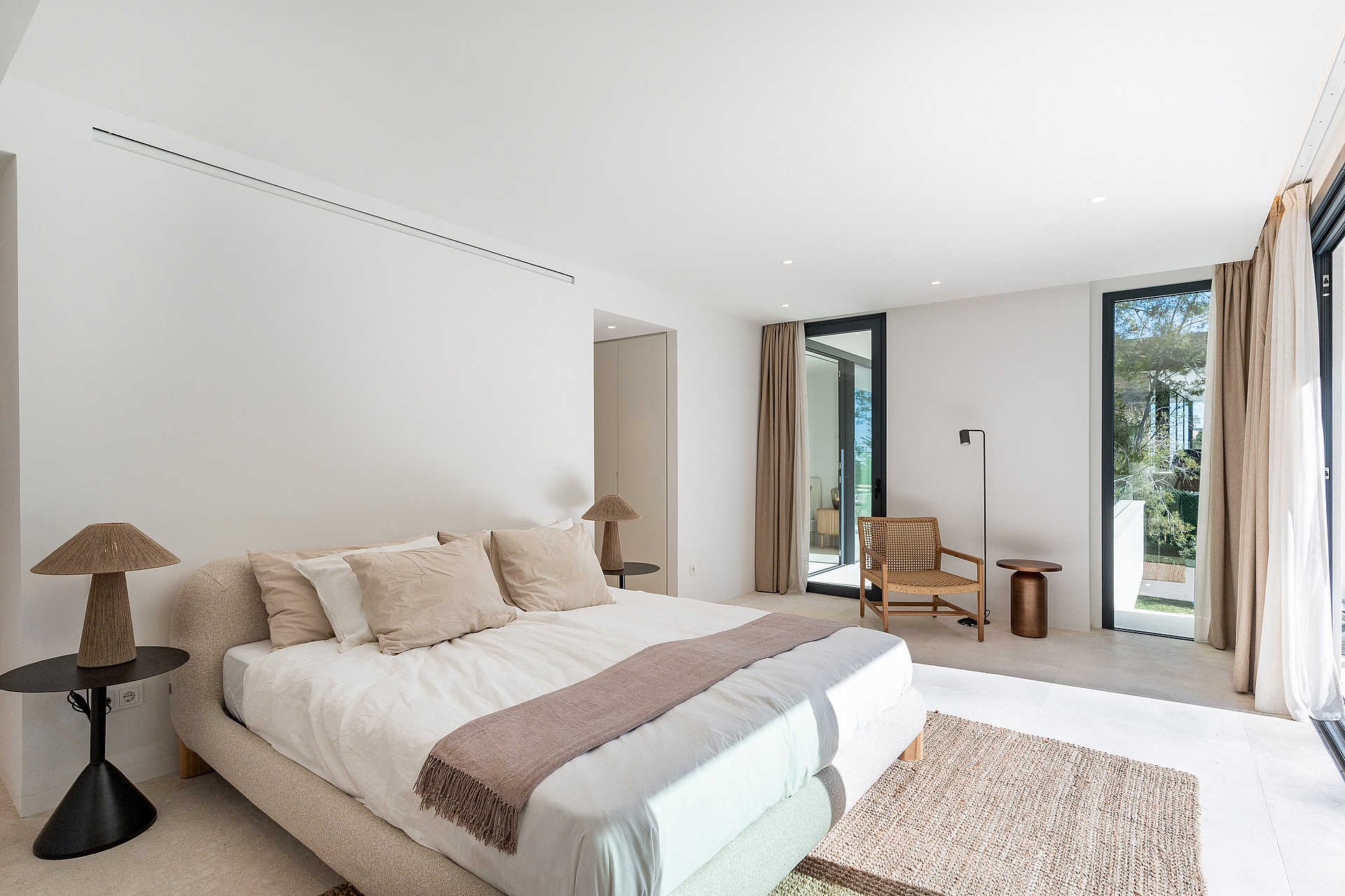 Serene master bedroom with neutral linen bedding, rattan accent chair and floor-to-ceiling sliding doors to a private terrace with pine tree views