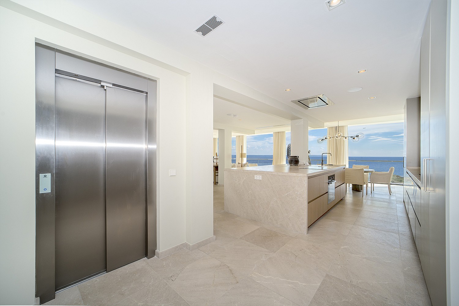 A modern kitchen interior featuring a brushed stainless steel elevator door, stone flooring, and an open layout facing the sea.