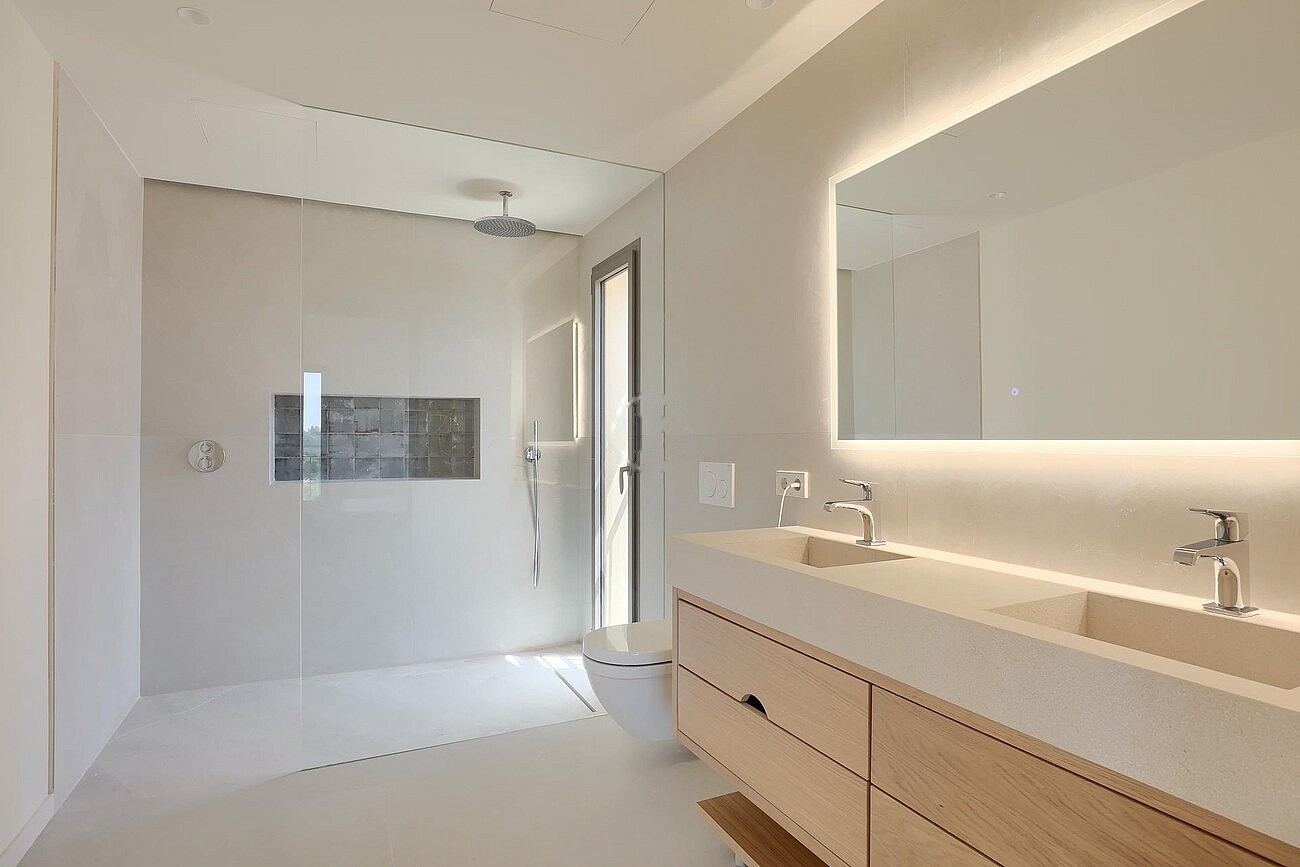 Bright bathroom with dual vanity, light wood cabinetry, walk-in shower with glass partition and wall-mounted rain showerhead.