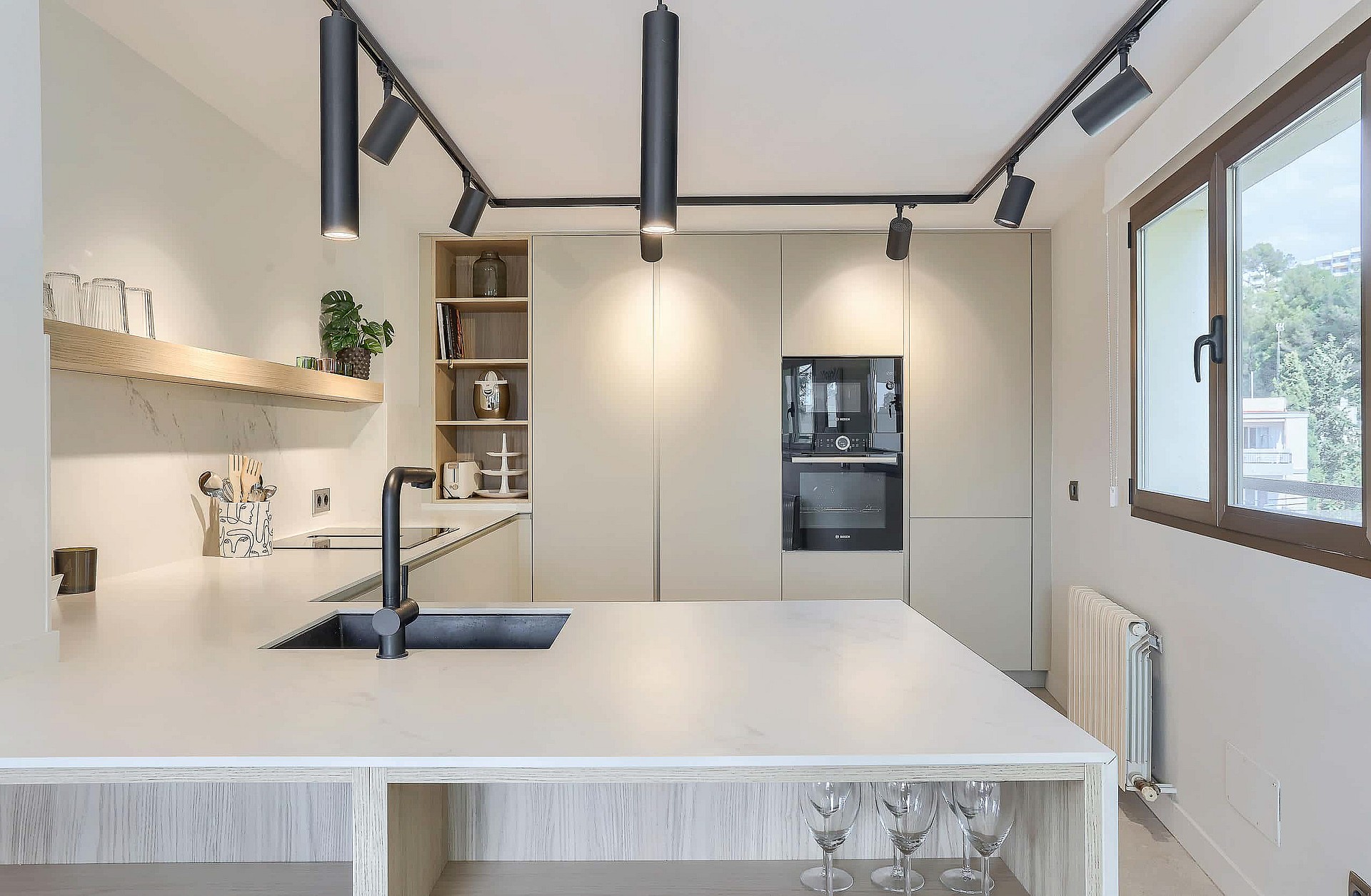 Modern L-shaped kitchen featuring matte white countertops, dark track lighting, and built-in wall oven in a neutral space.