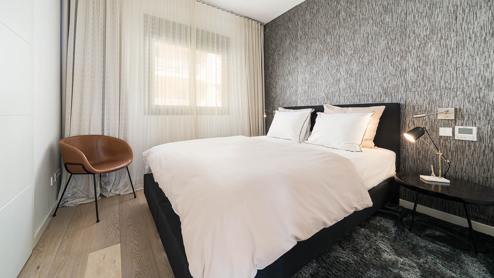 A modern bedroom with a black upholstered bed frame, light bedding, and a grey vertical-patterned accent wall. Sheer curtains cover the window to the left.