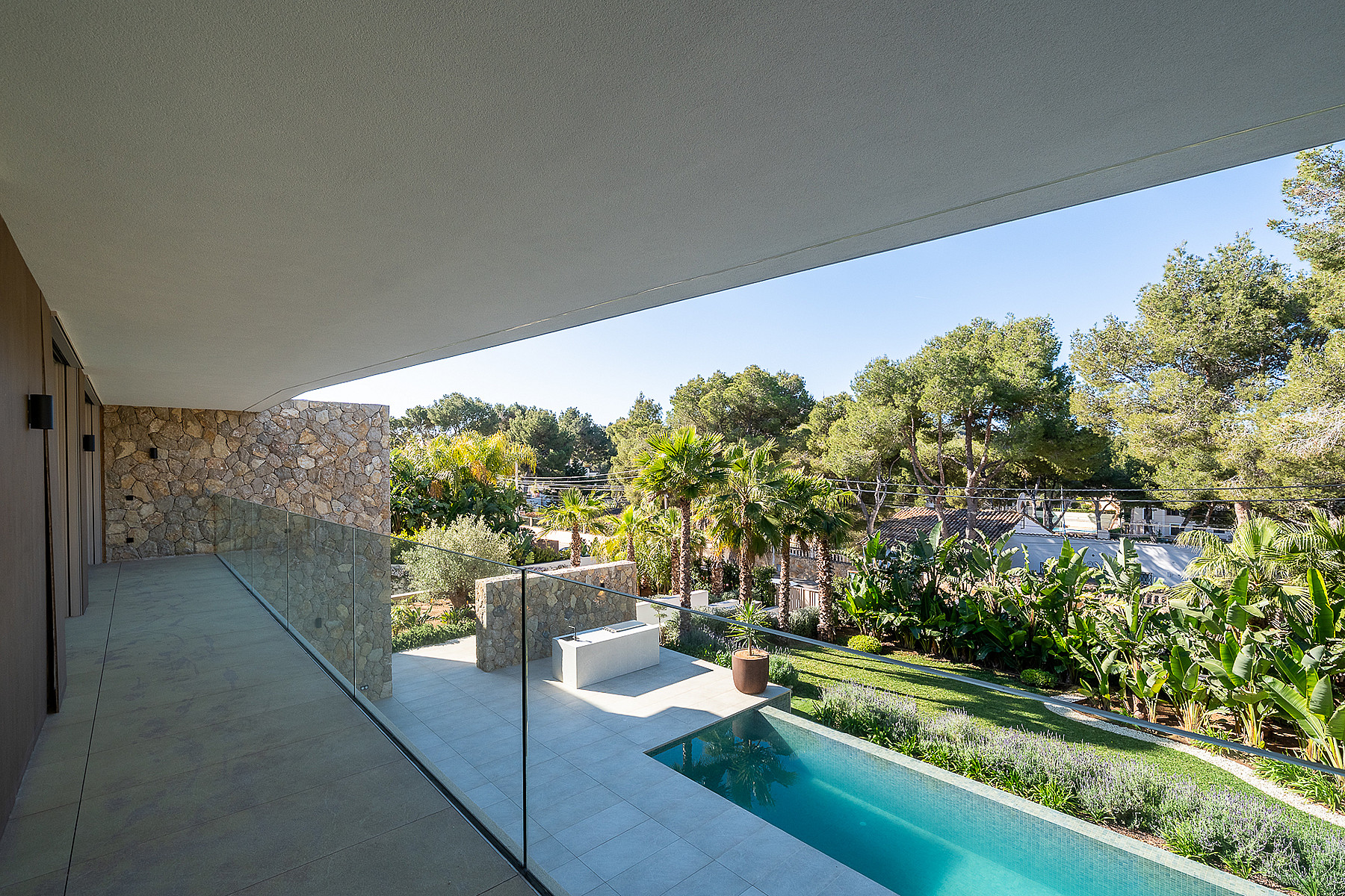 A wide, covered terrace with natural stone walls and a glass balustrade, overlooking a private garden with a swimming pool.