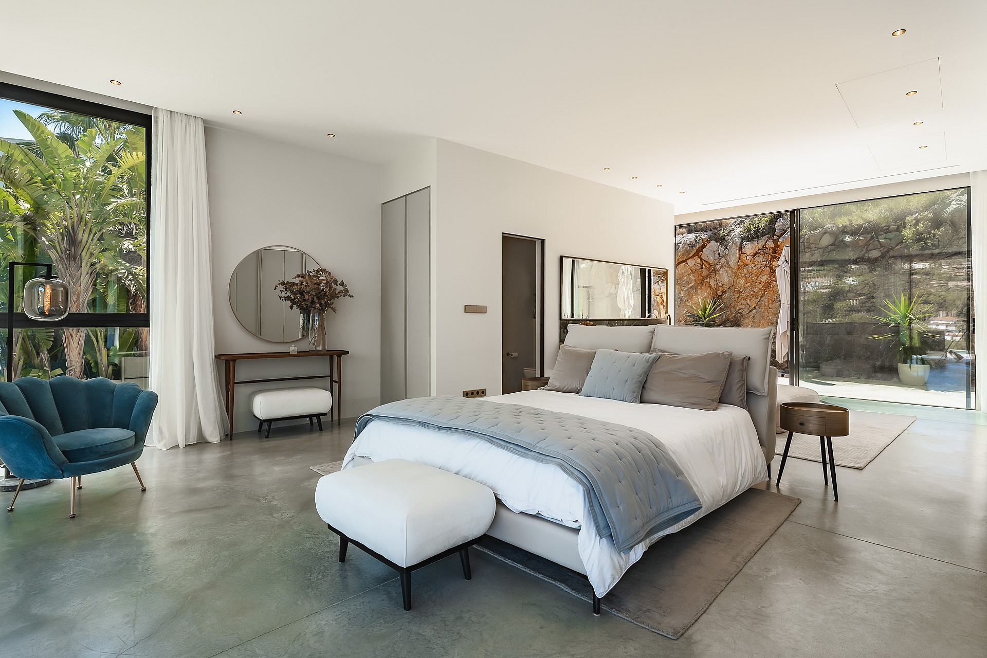 Modern master bedroom with polished concrete floors, floor-to-ceiling windows, a blue velvet chair, and grey bedding.