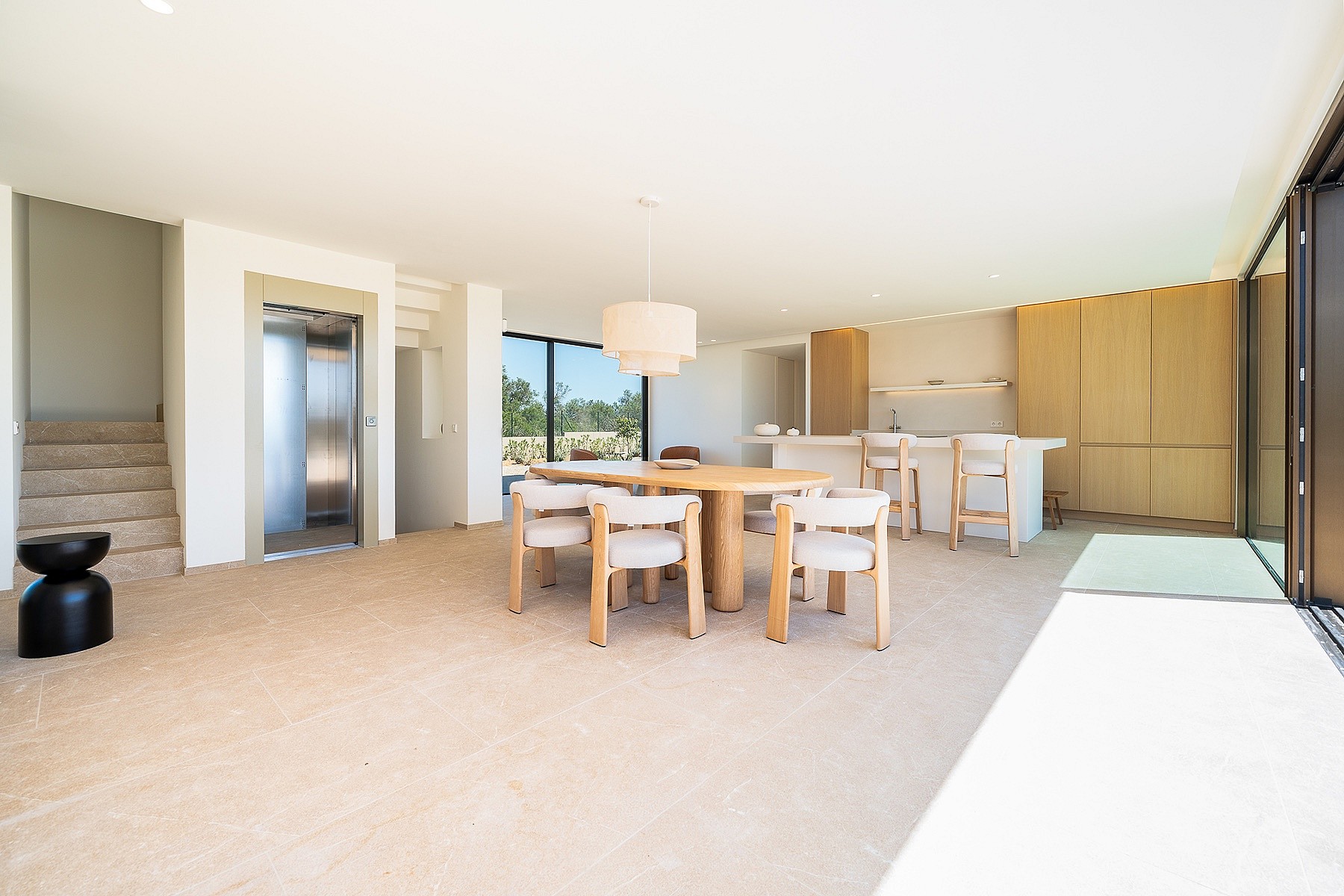 Interior view of an open-plan kitchen and dining area featuring a light wood dining table, cream chairs, a central home elevator, and floor-to-ceiling glass doors.