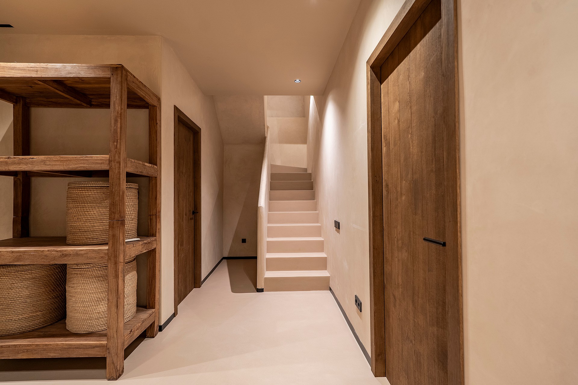 A neutral-toned hallway in a Genova townhouse featuring a wooden shelving unit with wicker baskets, solid wood doors, and a limestone staircase leading upwards under soft recessed lighting.