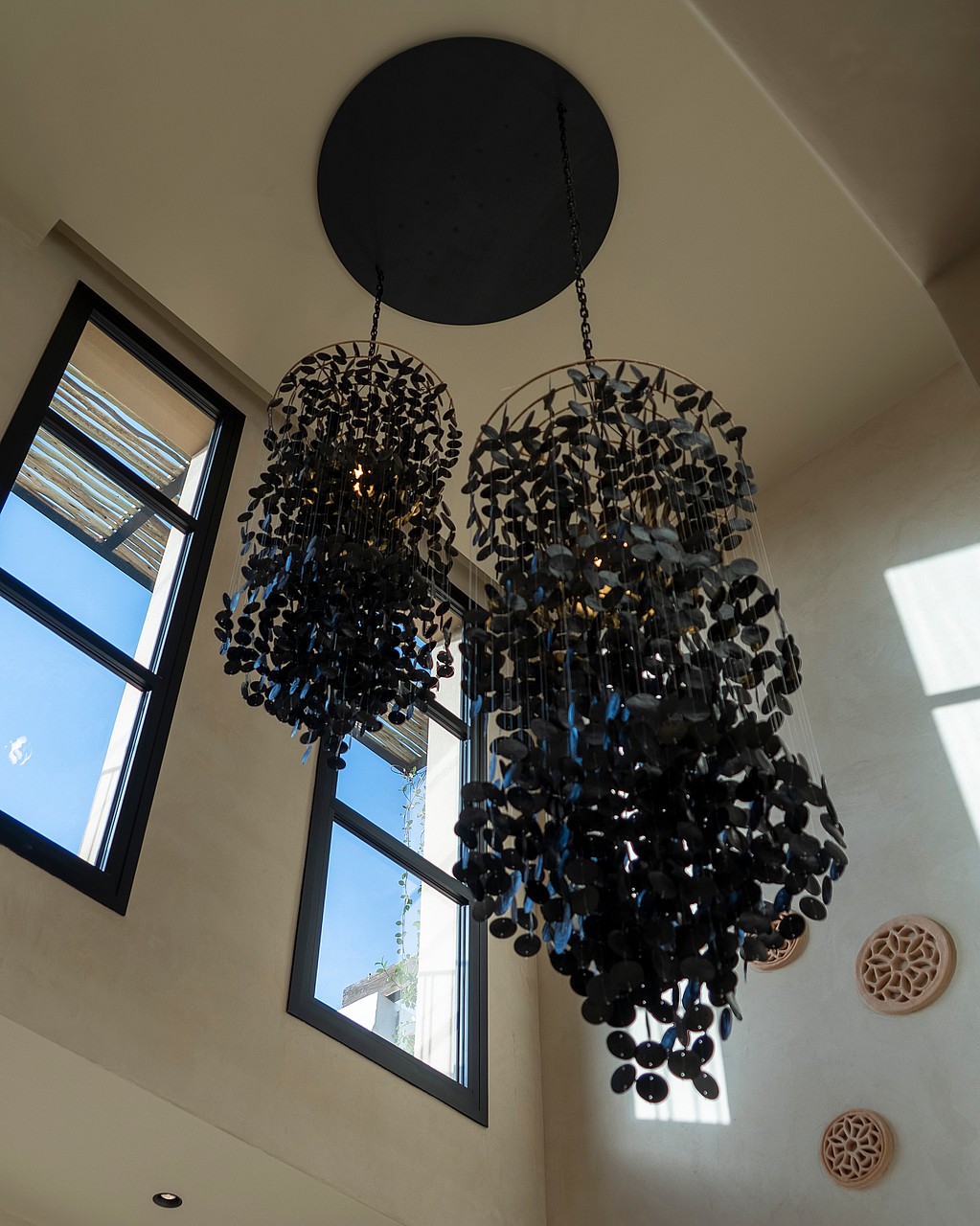 A low-angle shot of two dark, intricate, floral-inspired pendant light fixtures hanging from a high ceiling near tall windows.