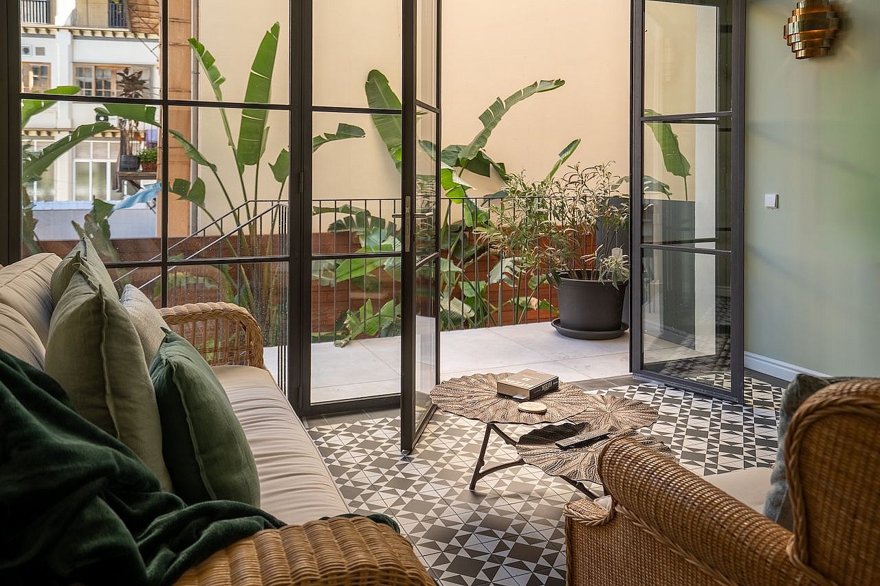 A living space with rattan furniture and patterned floor tiles opening through black-framed glass doors onto a green terrace.