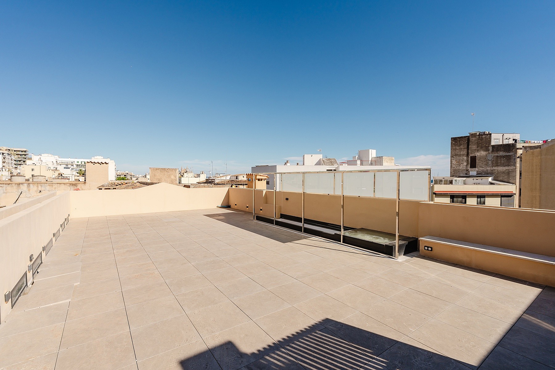 A wide, empty rooftop terrace paved with light tiles, featuring low perimeter walls, glass balustrades, and distant city buildings under a clear blue sky.