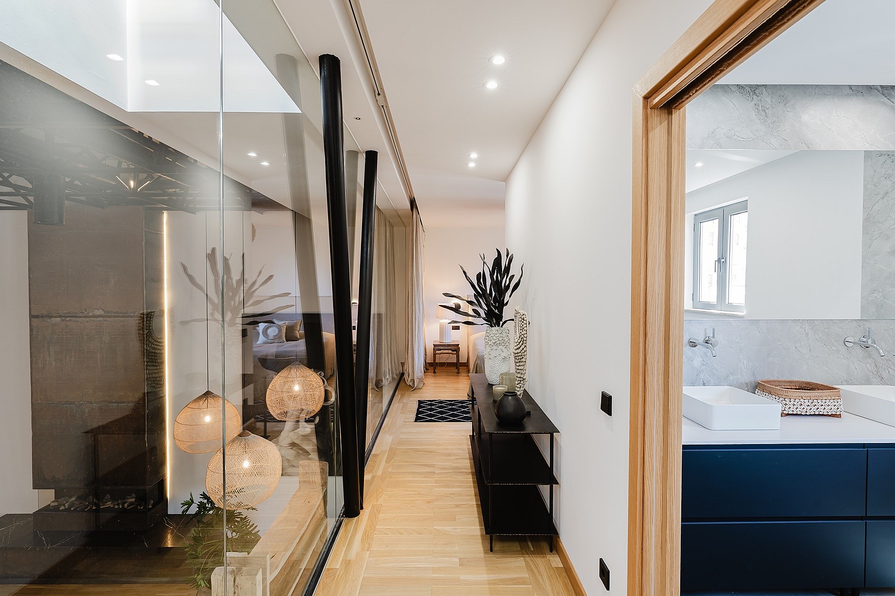 A bright hallway with light wood flooring leading to a master bathroom with a navy vanity, separated by a glass wall.