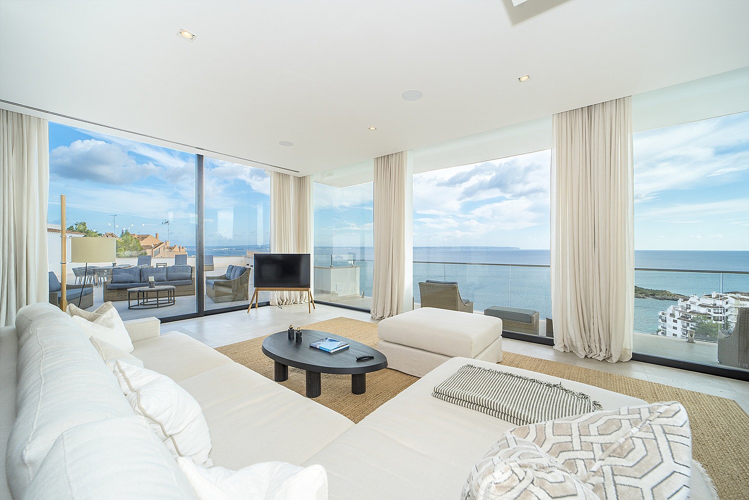 Modern living room featuring floor-to-ceiling glass walls, white sofas, woven area rug, and panoramic coastal views.