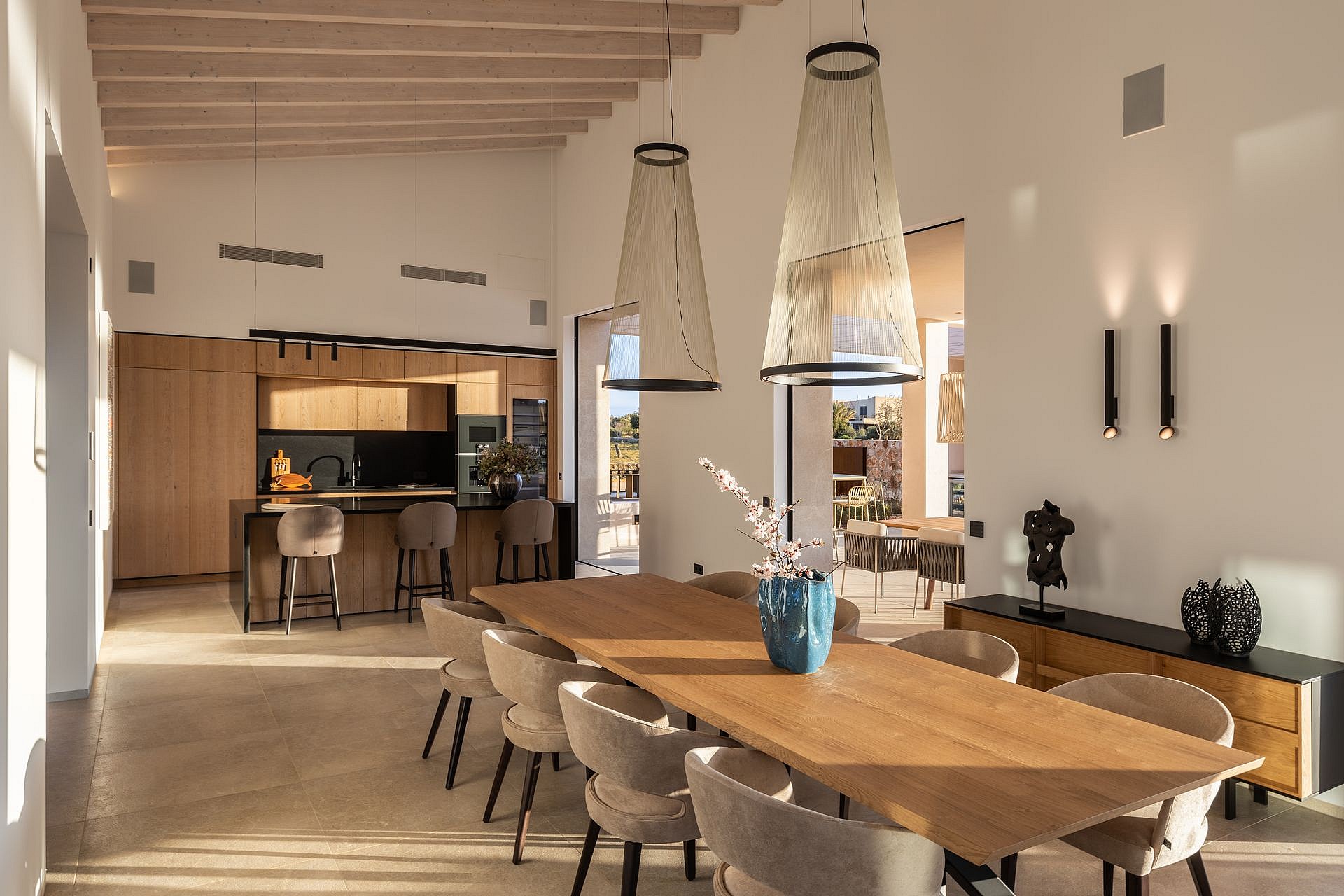 An open-concept interior featuring a long wooden dining table, modern mesh pendant lights, and a kitchen with light wood cabinetry and black accents.