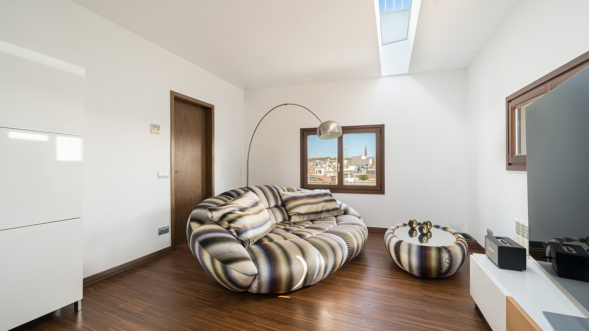 Modern living room with a striped round sofa, dark wood floors, a skylight, and a window viewing Palma Old Town.